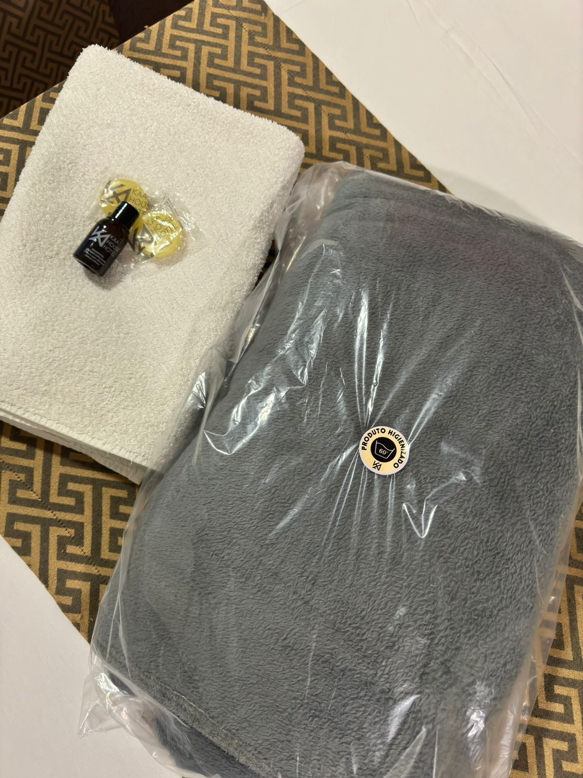 A gray towel and a bottle of lotion on a white towel, both in a plastic bag, on a patterned floor.
