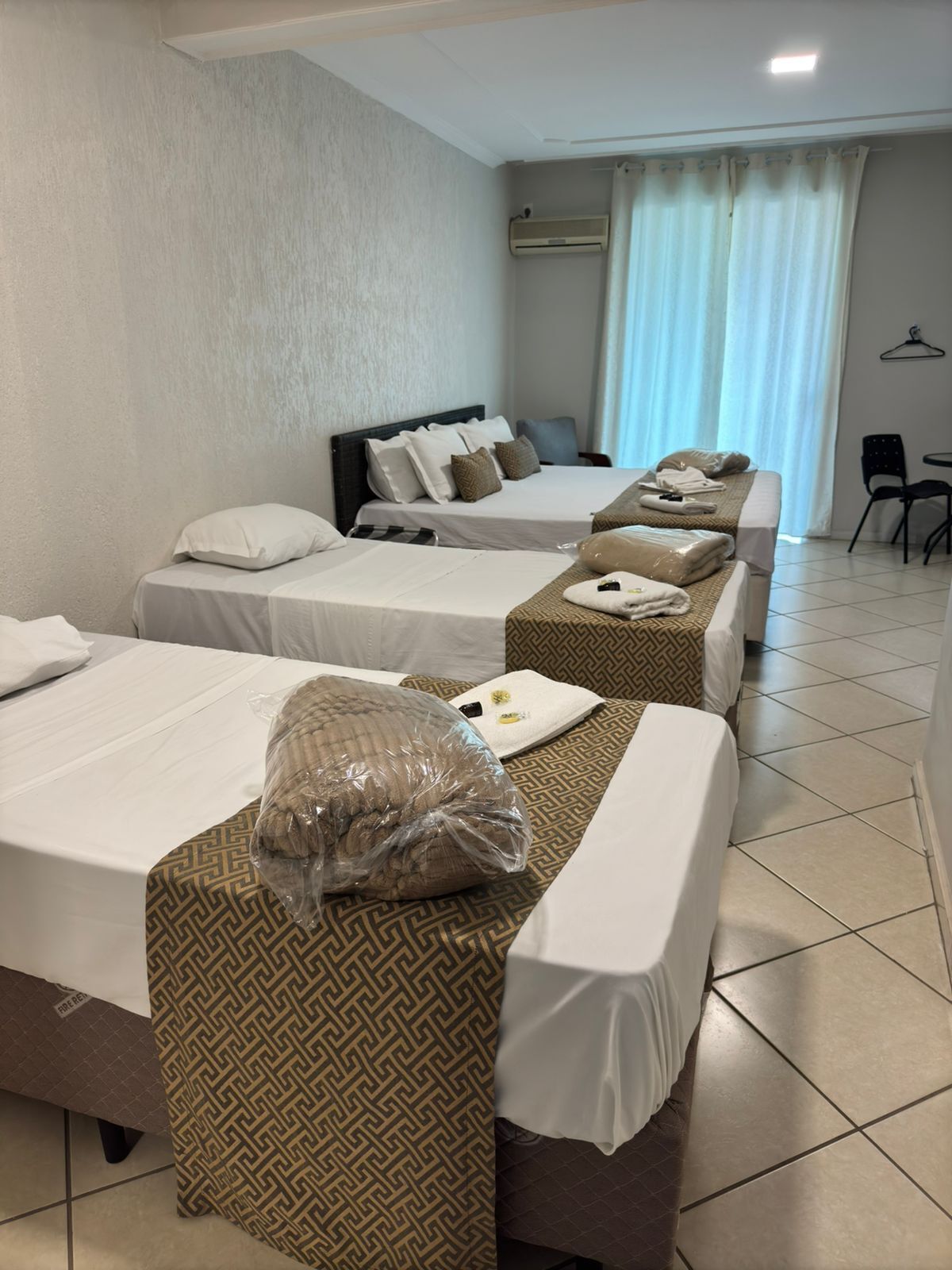 A hotel room with three beds, each with a patterned bedspread, a large pillow, and a blanket. There is a chair and a small table in the corner.