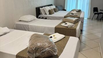 A hotel room with three beds, each with a patterned bedspread, a large pillow, and a blanket. There is a chair and a small table in the corner.
