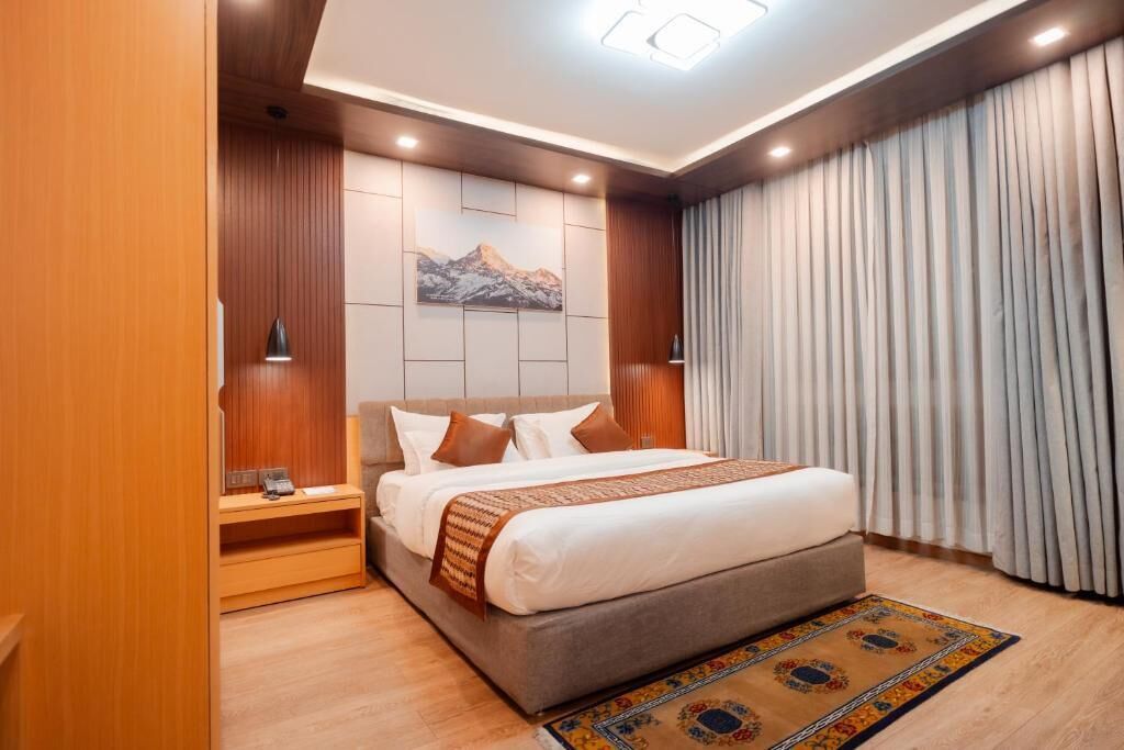 A modern hotel room with a large bed, wooden paneling, a mountain artwork on the wall, and a large window with curtains.