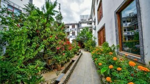 A narrow pathway flanked by lush greenery and vibrant flowers, leading to a building with large windows and a traditional architectural style.