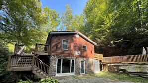 A rustic cabin with a stone foundation, wooden deck, and a small porch. - Charming riverfront home with beautiful views, hot tub and game room. Fishing! (Elkins)