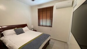 Ein Schlafzimmer mit einem Bett, Nachttisch, Lampe, Fenster mit Jalousien, Klimaanlage und einem Fernseher.
