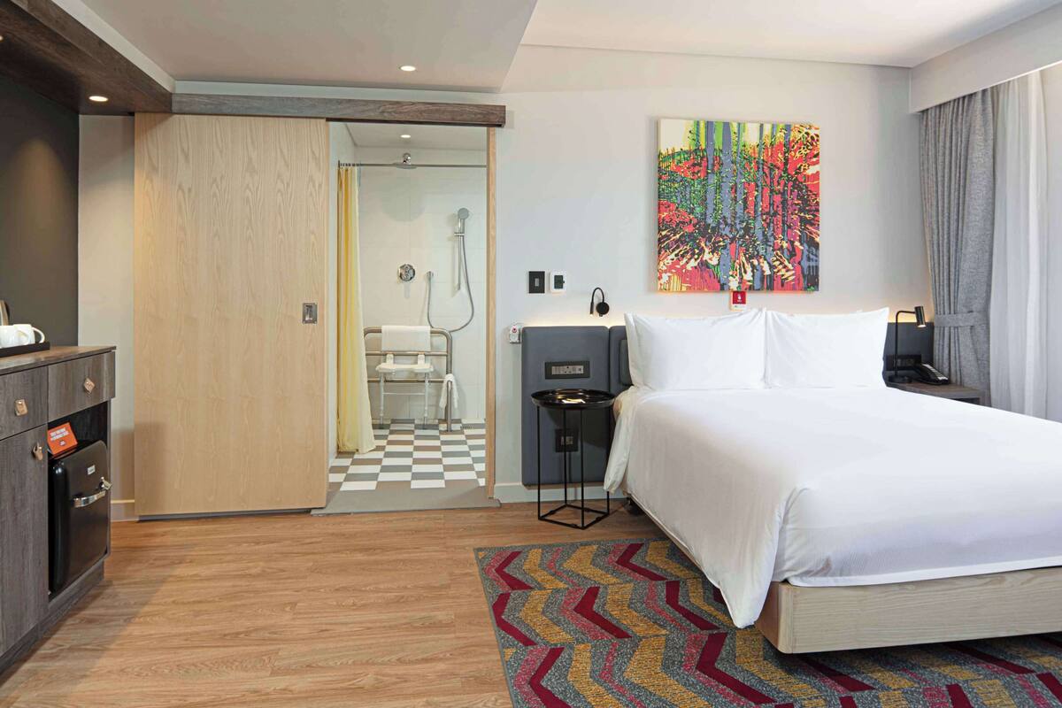 Habitación de hotel con cama, escritorio, silla, mesita pequeña, una obra de arte colgada en la pared y un baño visible a través de una puerta abierta.
