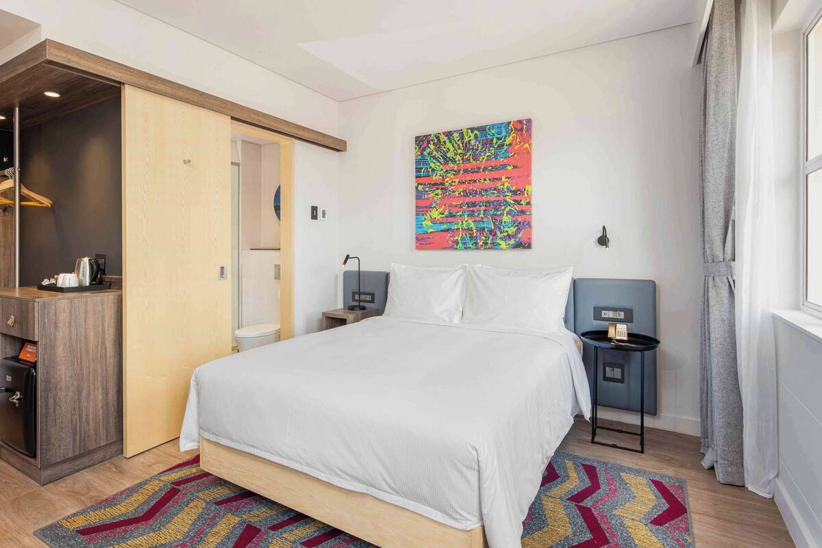Una habitación de hotel con cama, mesitas de noche, armario y un cuadro abstracto y colorido en la pared.