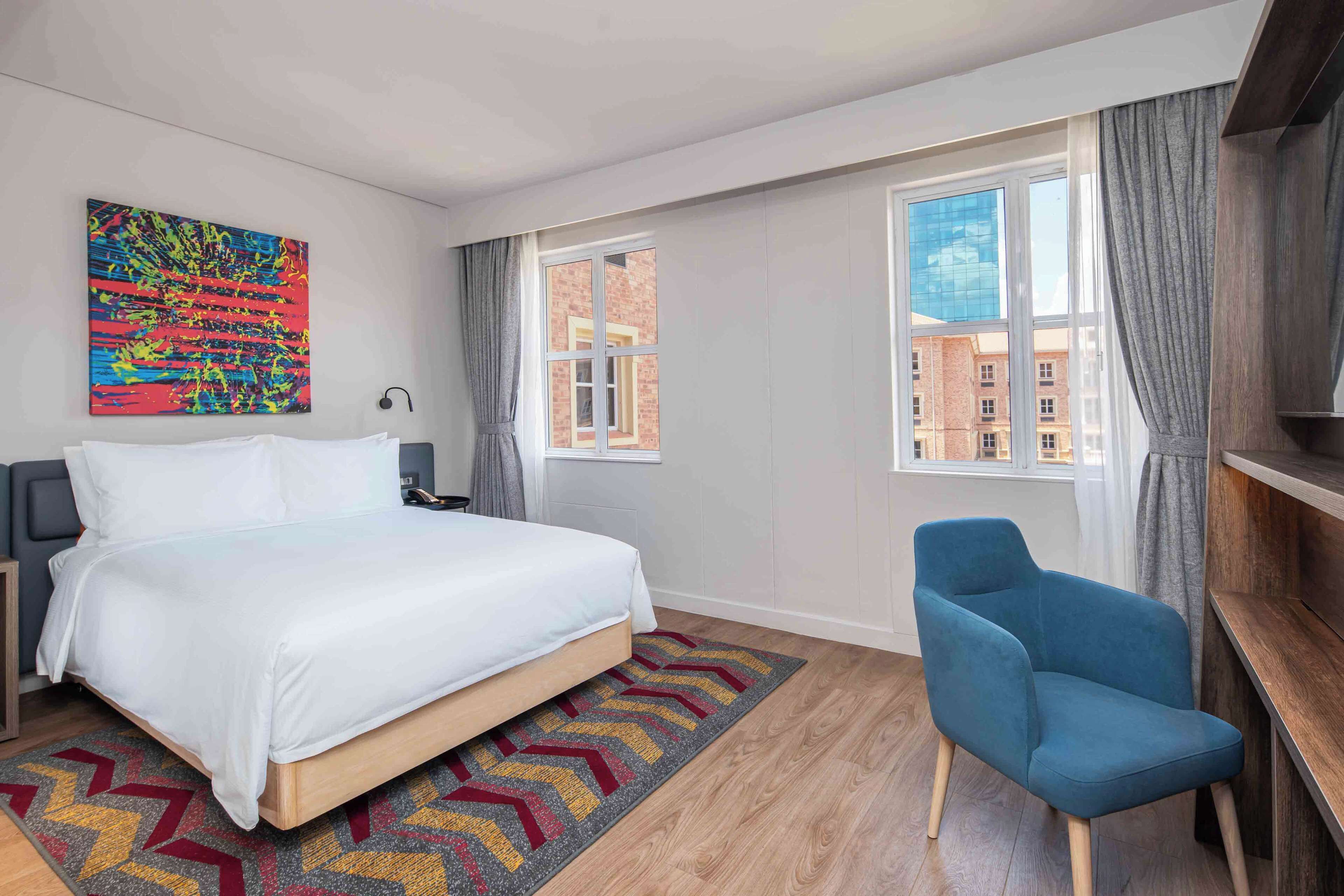 A hotel room with a bed, a blue armchair, a colorful abstract painting, and a window with a view of buildings.