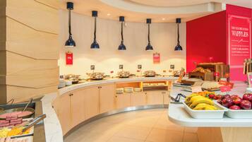 Un espace buffet proposant une variété de plats, notamment des fruits et un poste où l’on prépare des gaufres.