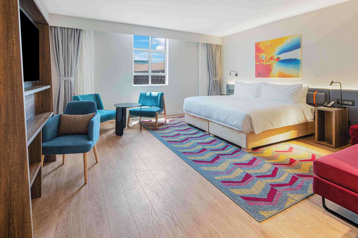 Una habitación de hotel con una cama, dos sillones azules, una mesita, un sofá rojo, una alfombra colorida y un cuadro en la pared.