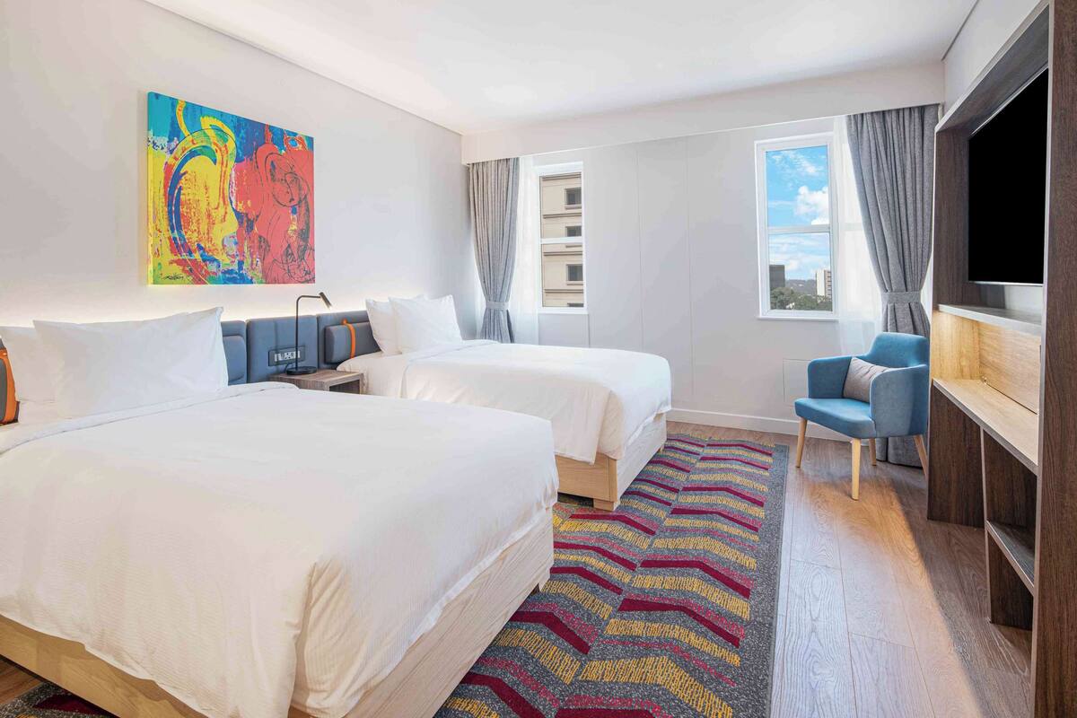 Habitación de hotel con dos camas, televisión, escritorio, silla, alfombra colorida y un cuadro en la pared.