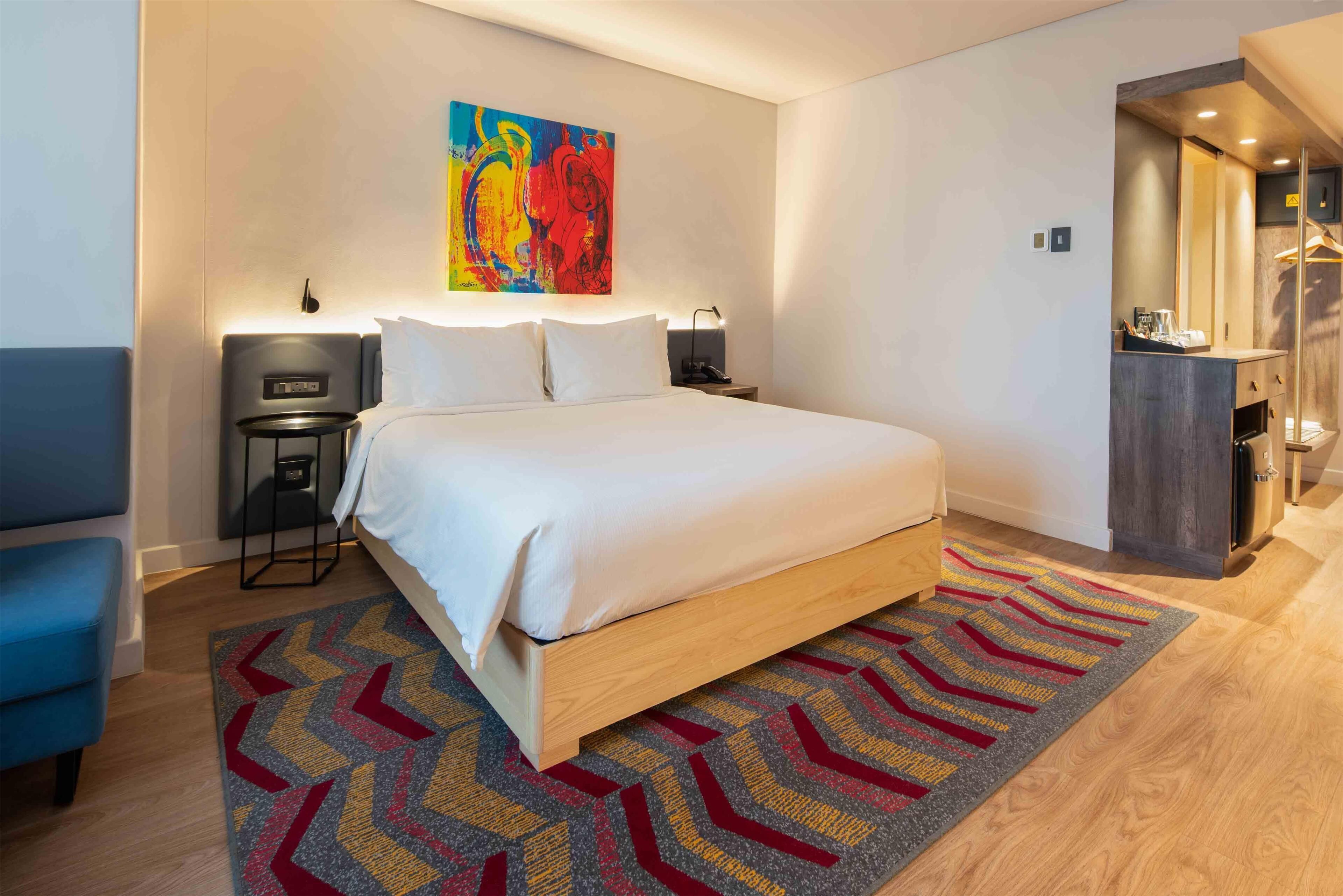 A modern hotel room with a large bed, a desk, a chair, a colorful abstract painting, and a walk-in closet.