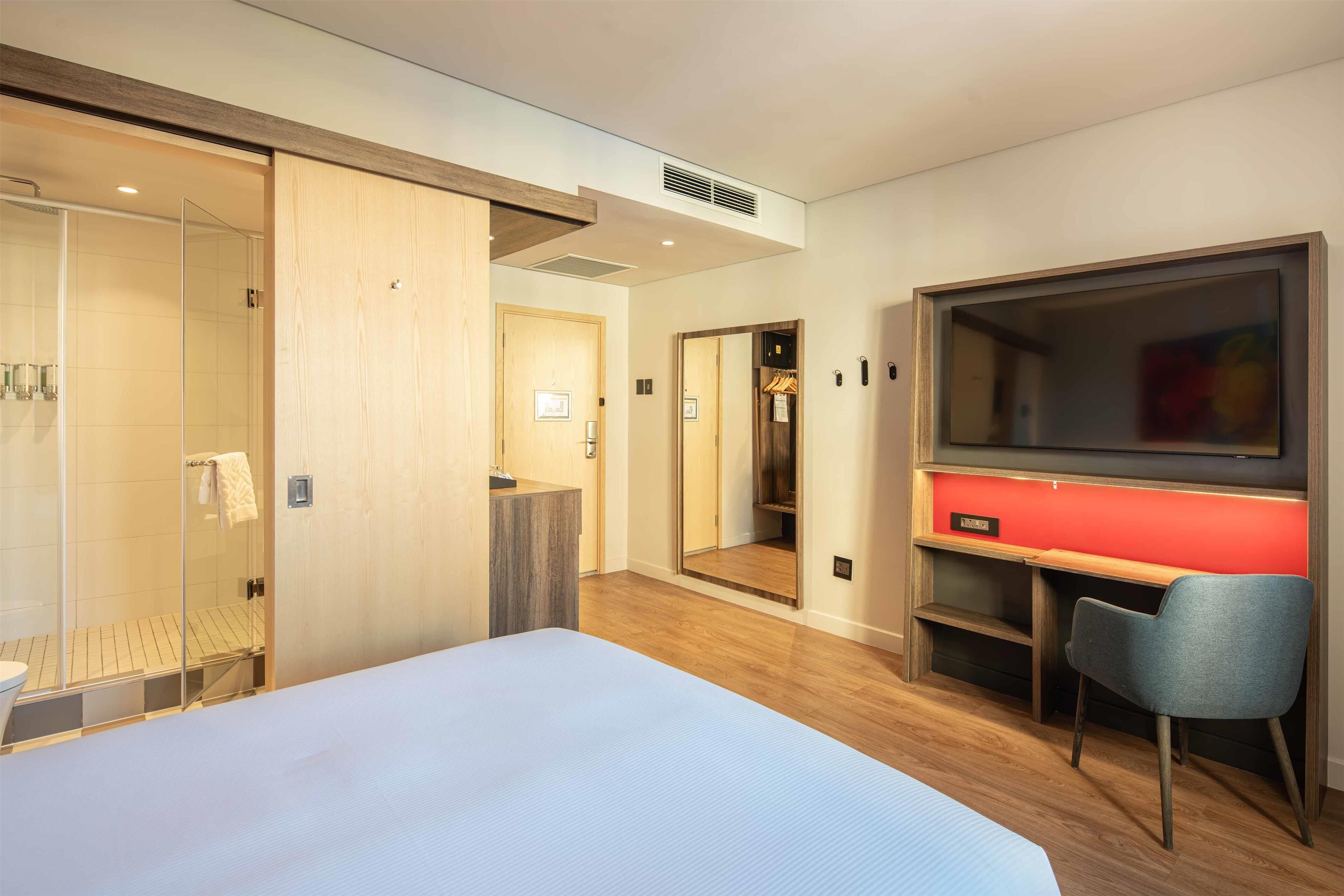 A modern hotel room with a large bed, a desk, a TV, a mirror, and a walk-in closet.