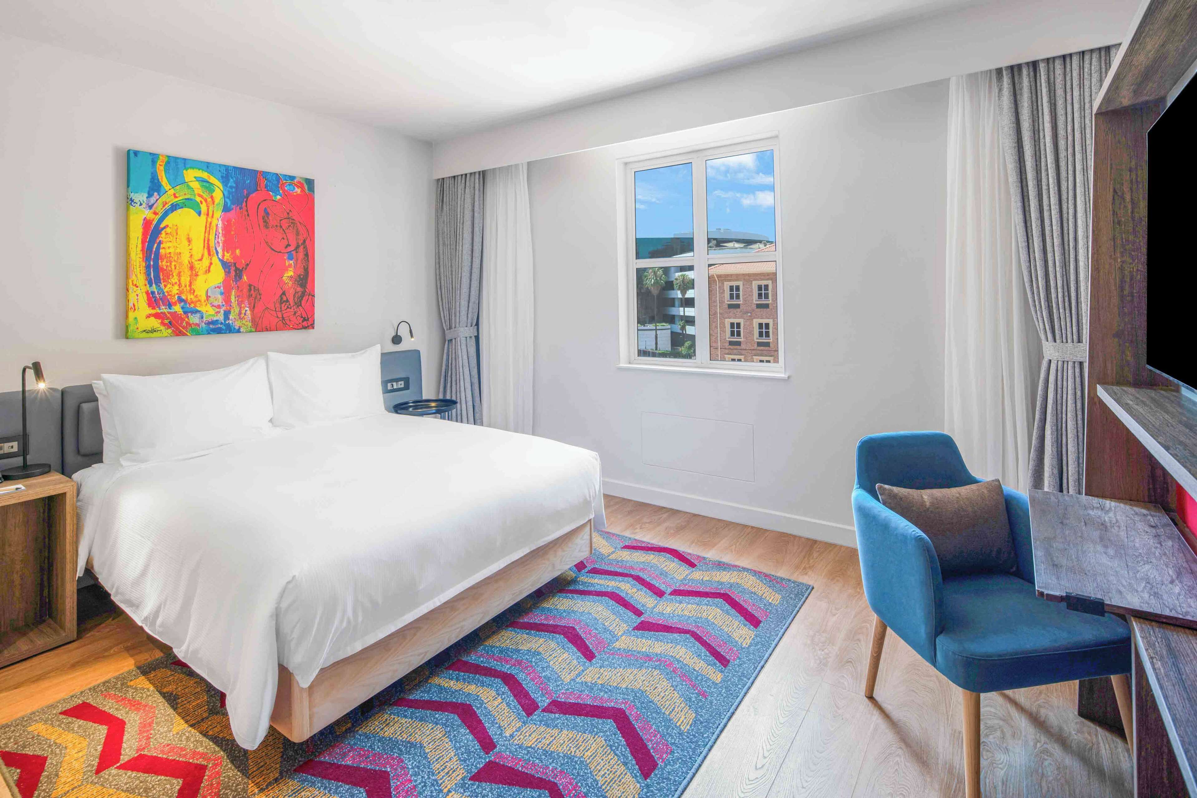 A hotel room with a bed, a desk, a chair, a colorful rug, a painting on the wall, and a window with a view of buildings.