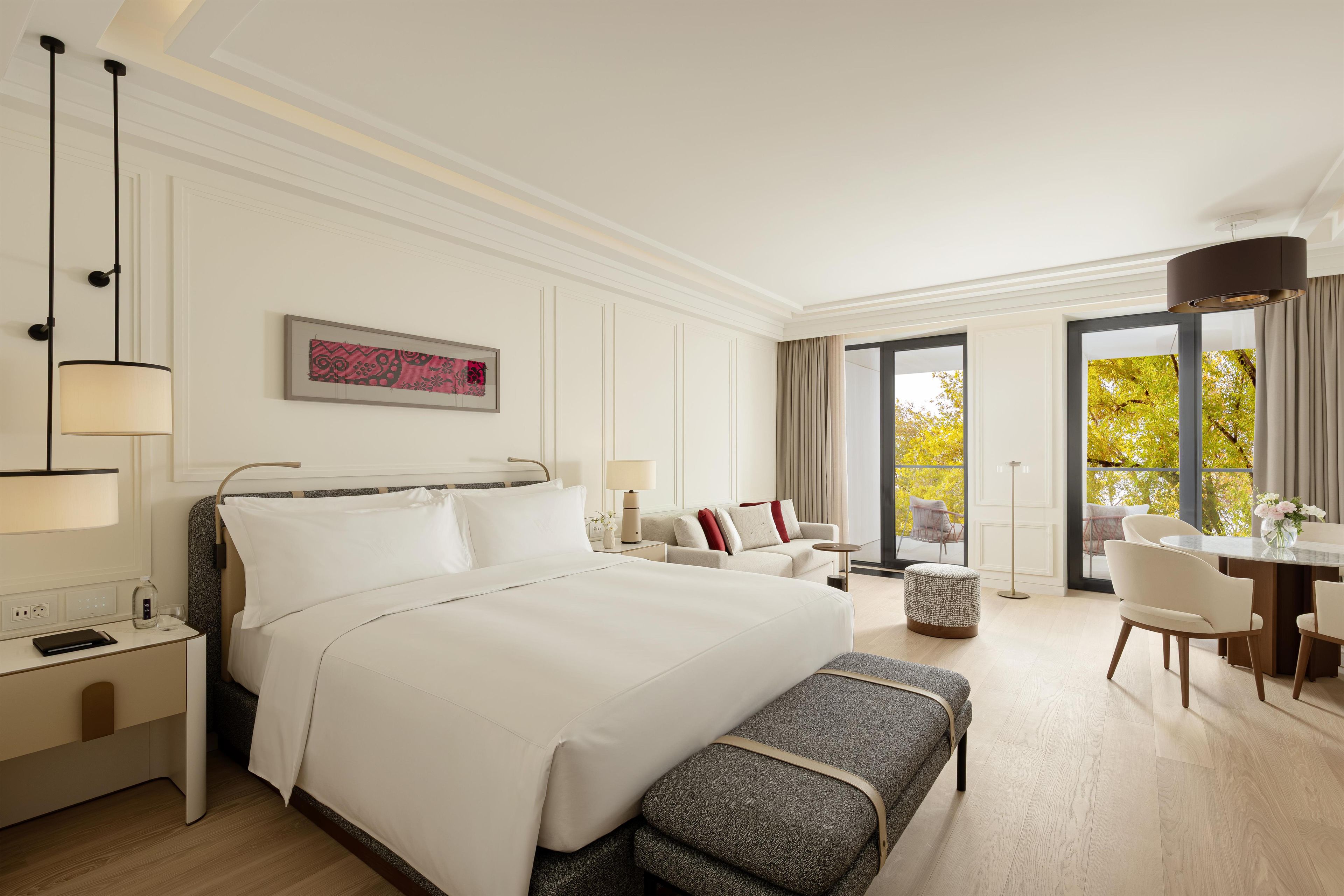 A hotel room with a large bed, bedside tables, a desk, and a view of trees through the window.