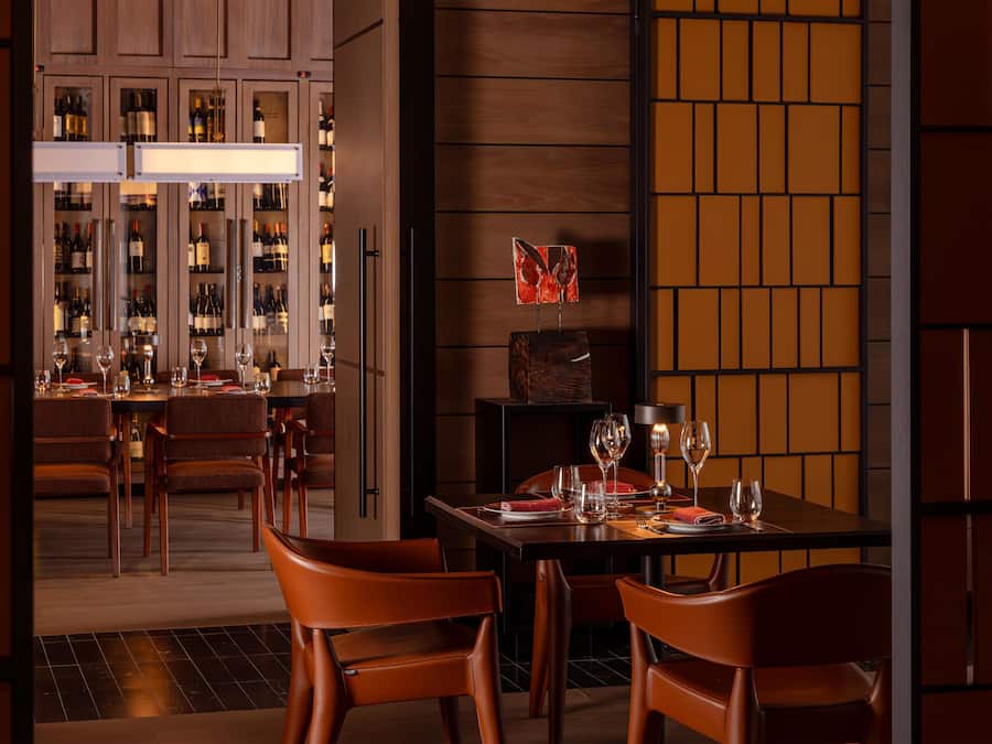 Interior de restaurante com área de bar visível através de uma divisória de vidro, com garrafas e copos de vinho.