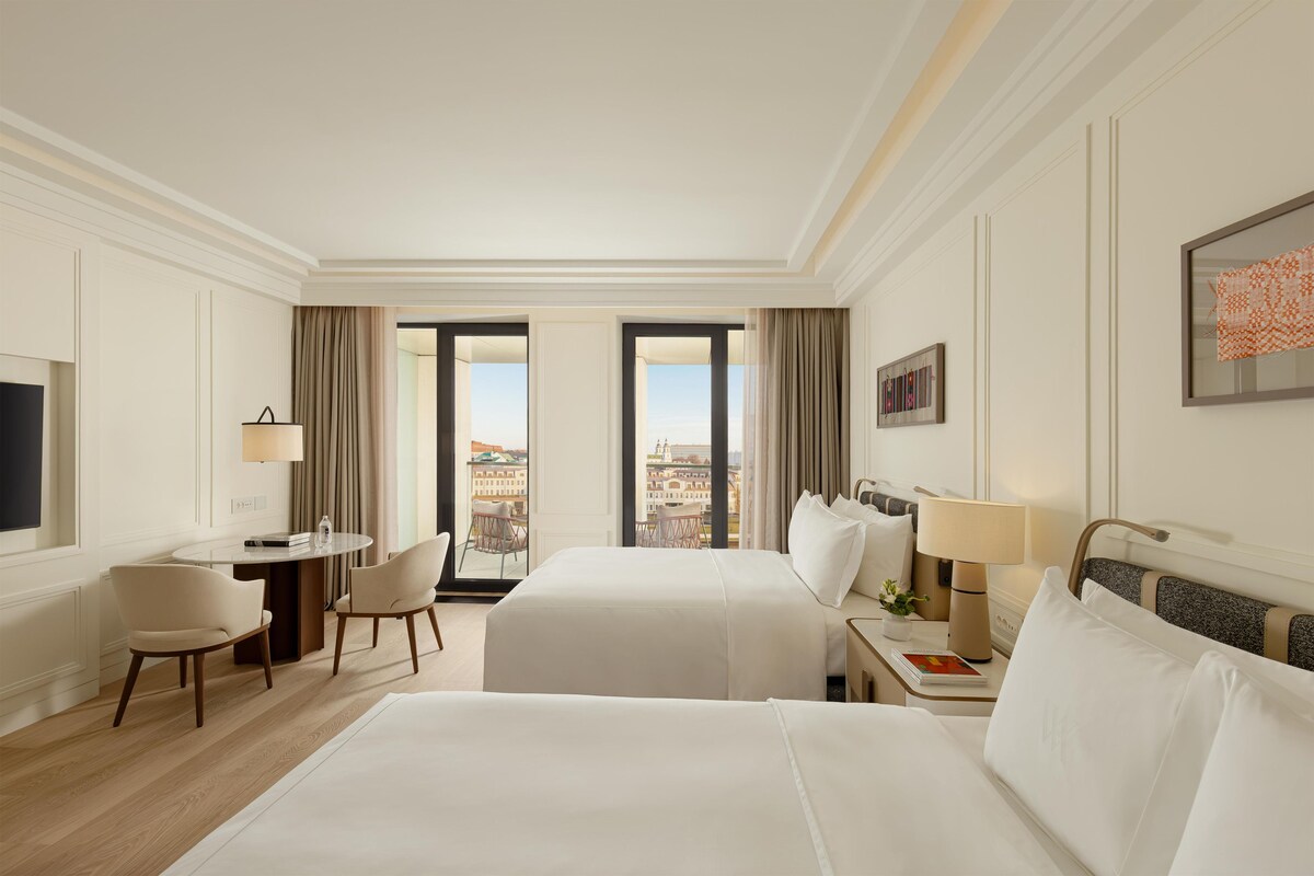 Een hotelkamer met twee bedden, een bureau, een stoel, een lamp en uitzicht op de stad.