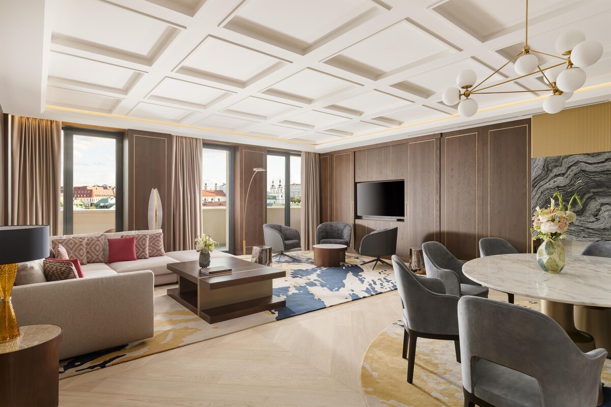 Een moderne hotelkamer met een grote flatscreen-tv, een bank, een eettafel met stoelen en uitzicht op de stad door grote ramen.
