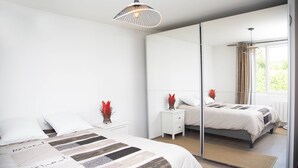 A bedroom with a large mirror, a bed with a patterned bedspread, a bedside table with a vase, and a window with curtains.