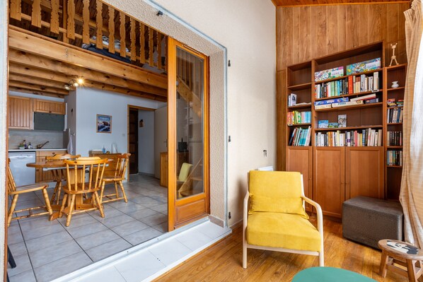 A cozy living room with a wooden bookshelf, a yellow armchair, and a dining area with wooden furniture.