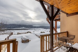 A wooden deck with a table and chairs, overlooking a snowy landscape with mountains in the distance.