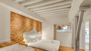 Un dormitorio con pared de ladrillo, suelo de madera, cama blanca y techo con vigas a la vista.