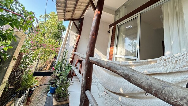A covered outdoor area with a wooden railing, potted plants, and a building with a large window. - Apartamento 2 Praia Morro de São Paulo  (Cairu)