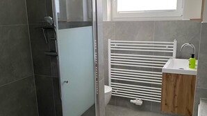 A modern bathroom with a glass shower enclosure, a toilet, a sink with a wooden vanity, and a window providing natural light. - Renovated vacation home in a quiet location on the outskirts of Berlin (Falkensee)