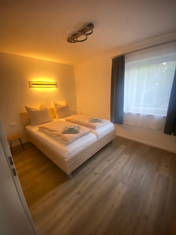 A bedroom with a bed, bedside lamps, a window with curtains, and a wooden floor.