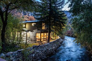 A rustic cabin with a stone wall and a wooden deck, overlooking a flowing river and surrounded by dense forest. - Cabin on Big Cottonwood River (Salt Lake City)