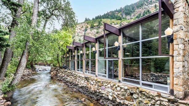Une rangée de cabanes aux murs de pierre, dotées de grandes fenêtres donnant sur un ruisseau et une colline boisée.