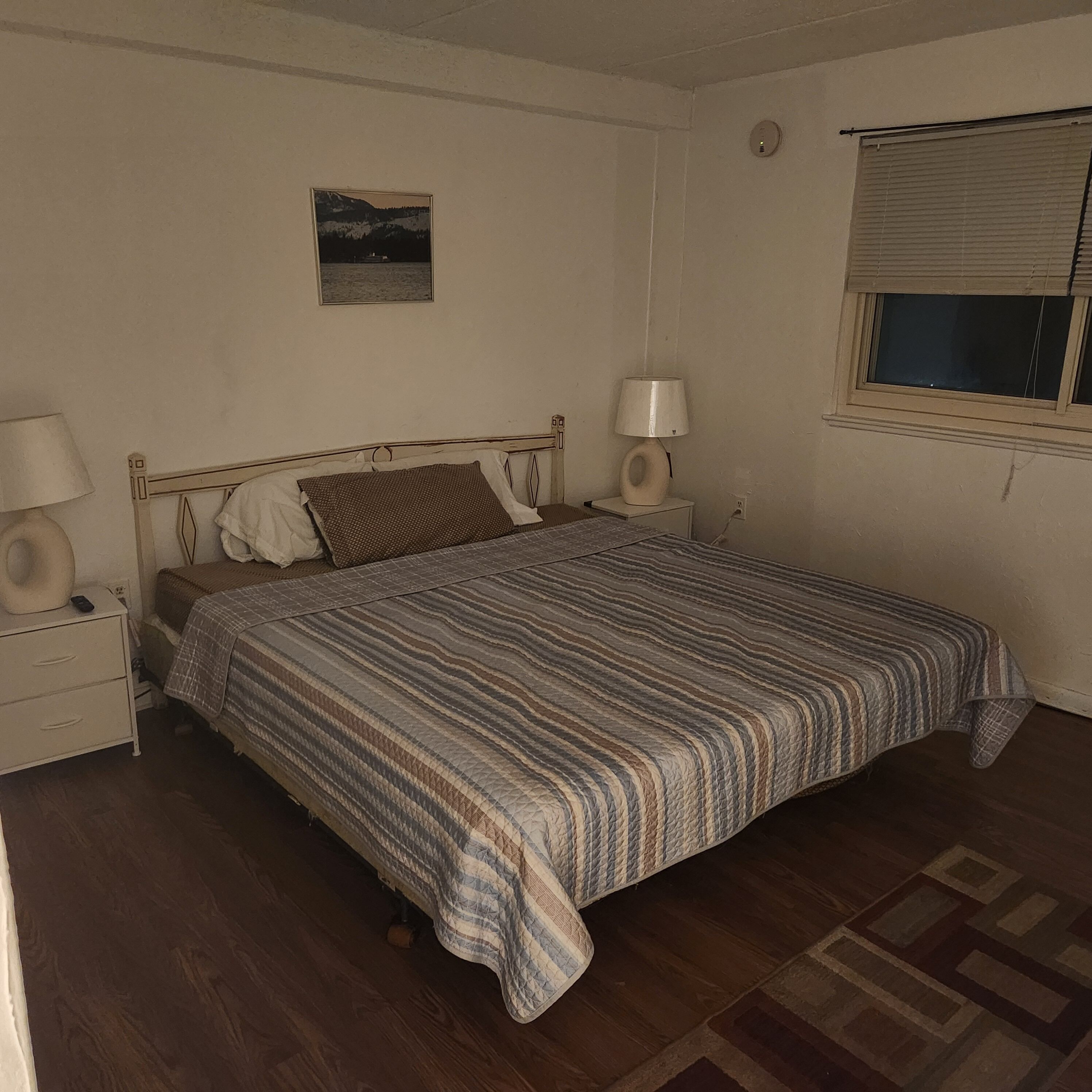 Une chambre à coucher avec un lit, deux tables de chevet, une commode, une lampe, un tableau au mur et une fenêtre avec des stores.