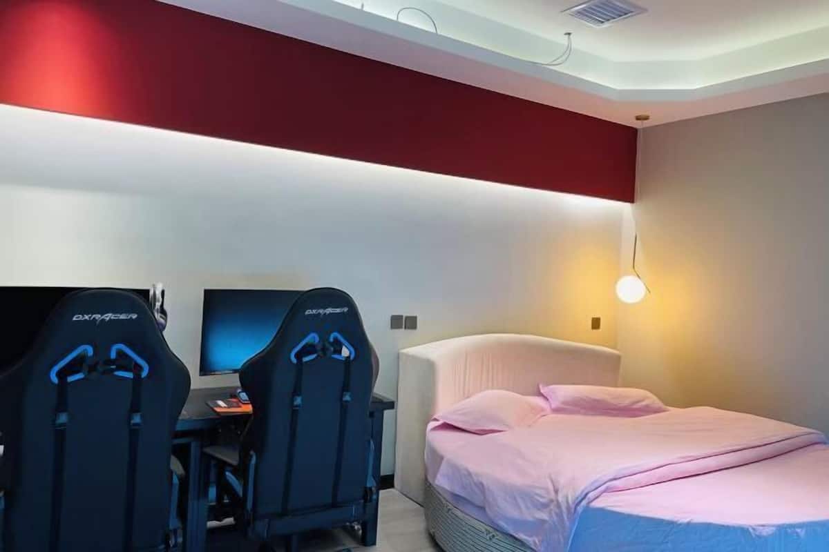 Een moderne hotelkamer met een bed, twee gaming stoelen, een bureau met een computer en een televisie.