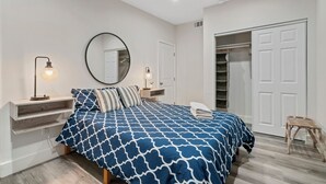 Ein Schlafzimmer mit Bett, Nachttischen, einem Spiegel, einem Kleiderschrank und einem kleinen Hocker.
