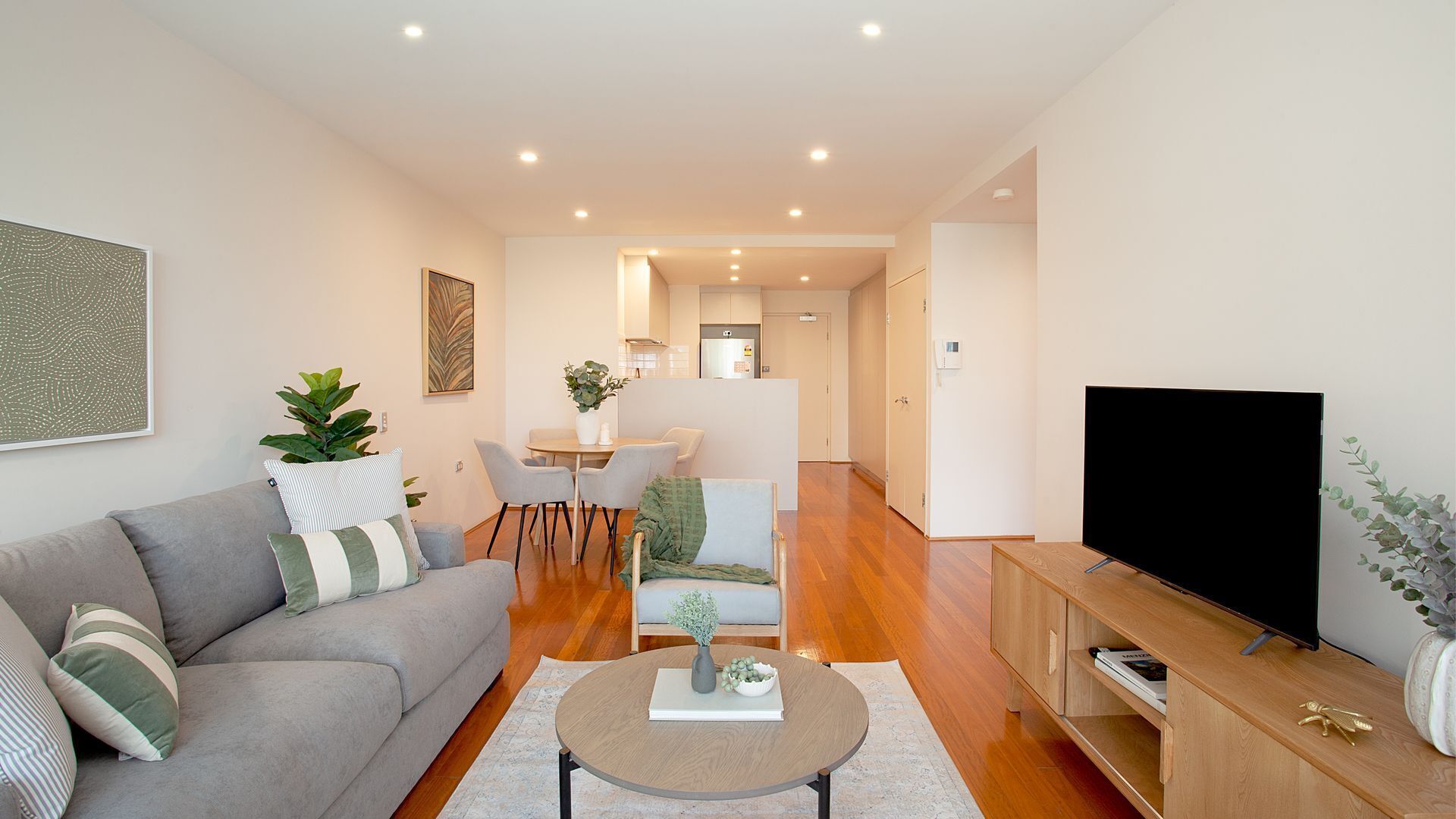 A modern living room with a grey sofa, a round coffee table, a flat-screen TV, and a dining area with a table and chairs.
