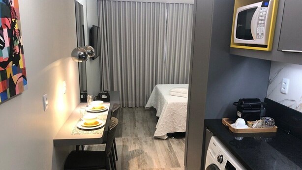 A compact hotel room with a dining area, a bed with a white bedspread, a washing machine, and a microwave on the countertop. - Apartment Asa Norte CLN 110 ap 111 (Brasília)