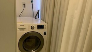 A compact, white washing machine in a small laundry room with a tiled floor and a curtain on one side. - Loft 34m² Asa Norte CLN 213 (Brasília)