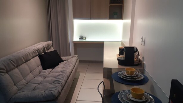 A compact living space with a sofa, a small table with cups, a coffee machine, and a window with blinds. - Apartment Asa Sul Golden Place (Brasília)