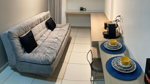 A compact living space with a sofa, a small dining table with plates and cups, a cabinet with shelves, and a curtain. - Apartment Asa Sul Golden Place (Brasília)