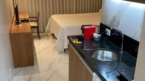 Habitación de hotel compacta con kitchenette, una cama con edredón blanco, un escritorio de madera y una silla.