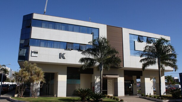 A modern building with a prominent 'K' logo, large windows, and a landscaped entrance area with palm trees.