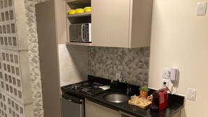 Cocina compacta con estufa a gas, lavamanos y microondas. Hay armarios sobre la encimera y una pequeña repisa con recipientes amarillos.