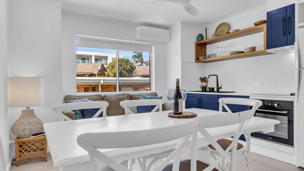 Eine moderne Küche mit weißen Schränken, ein Essbereich mit weißem Tisch und Stühlen sowie ein Blick auf die Nachbarschaft durch große Fenster.