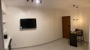Ein geräumiges Zimmer mit einem an der Wand befestigten Flachbildfernseher, einem Essbereich mit Tisch und Stühlen sowie einer Tür zu einem weiteren Raum.