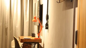 Een kleine kamer met een houten bureau, een stoel, een klein tafeltje, een hanglamp en een kapstok.