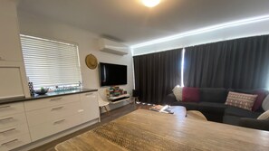 A modern living room with a flat-screen TV, a dark-colored sofa with cushions, a wooden coffee table, and a kitchen area with white cabinets and a window with blinds. - Alpine Mountain View 3 - Jindabyne (Jindabyne)