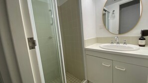 A bathroom with a glass-enclosed shower, a round mirror, a sink with two faucets, and a bottle of hand soap on the counter.