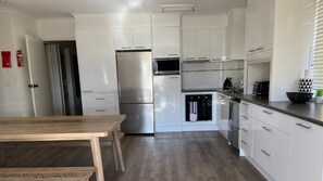 A modern kitchen with white cabinetry, stainless steel appliances, and a wooden dining table. - Alpine Mountain View 3 - Jindabyne (Jindabyne)