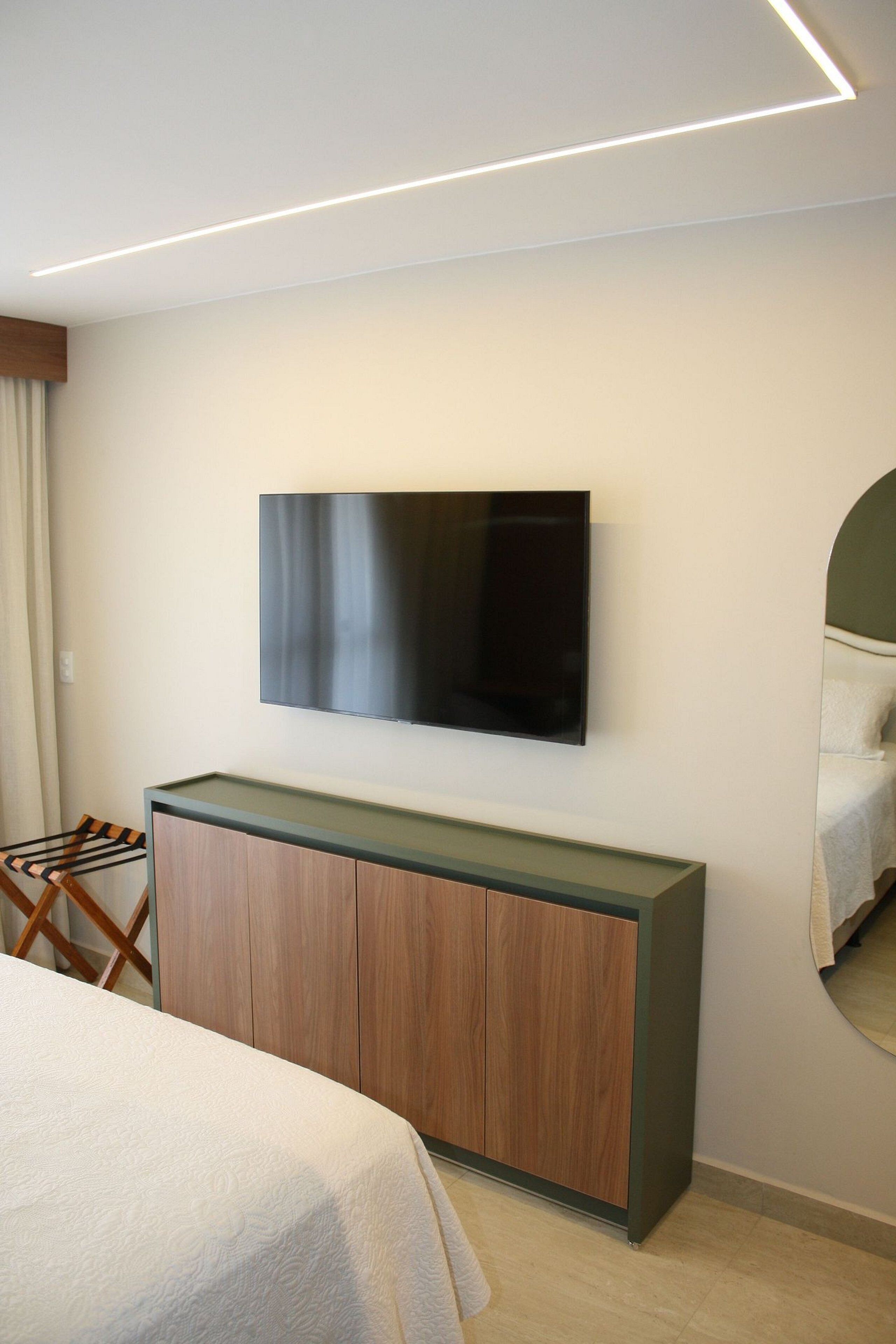 A modern hotel room with a flat-screen TV mounted on a wooden console, a bed with a white duvet, and a large mirror reflecting the bed.