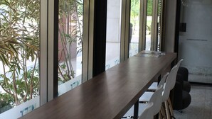 Une longue table en bois, des chaises blanches, des lampes suspendues et de grandes fenêtres ornées de plantes.