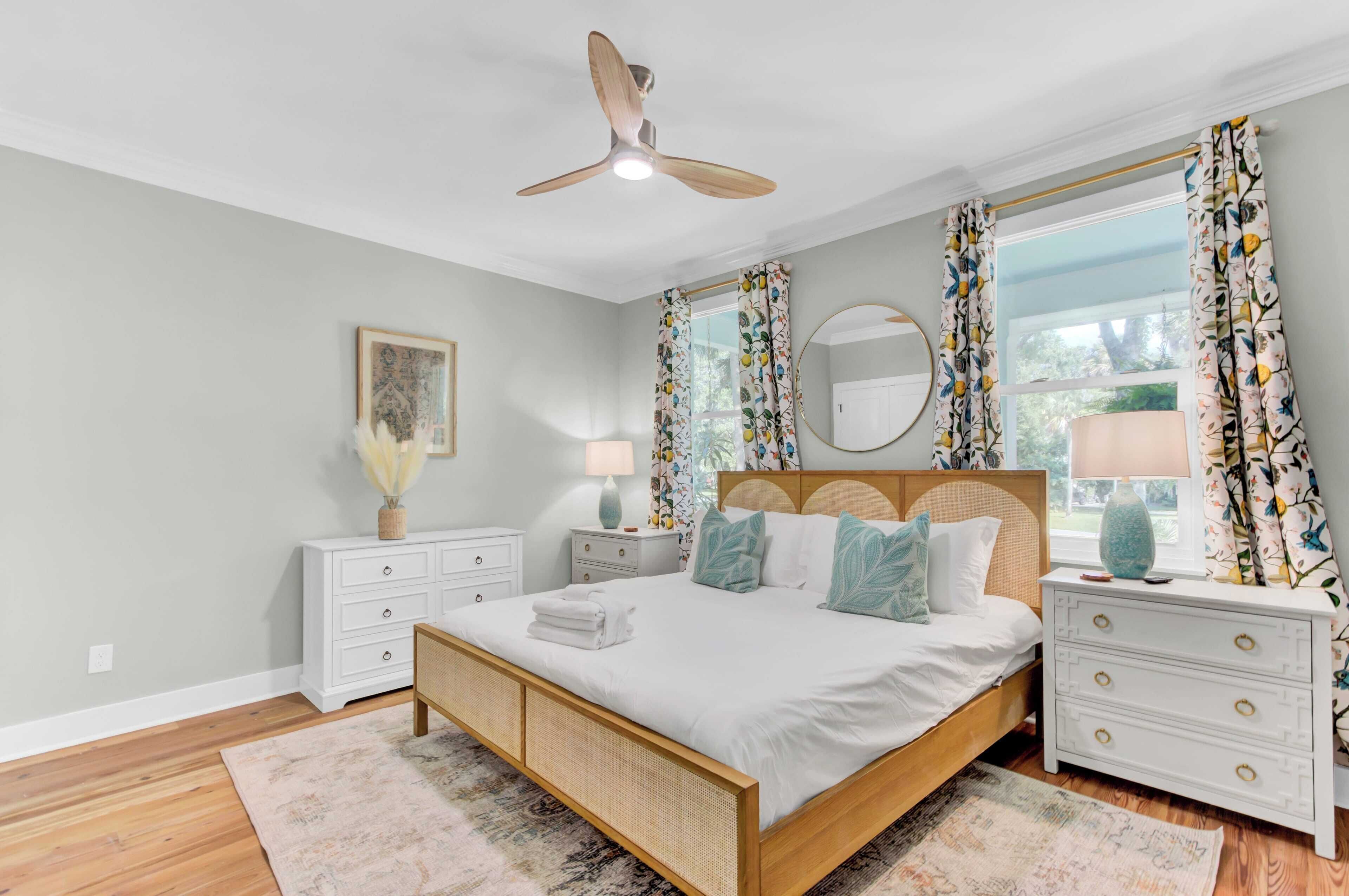 Une chambre à coucher avec un lit en bois, des tables de chevet blanches, un ventilateur de plafond et une grande fenêtre ornée de rideaux à motifs floraux.