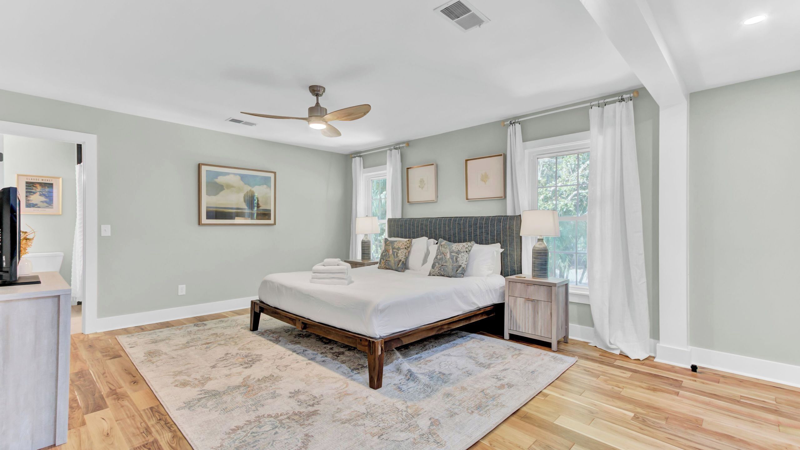 A bedroom with a bed, a ceiling fan, a window with curtains, a bedside table with a lamp, and a rug.